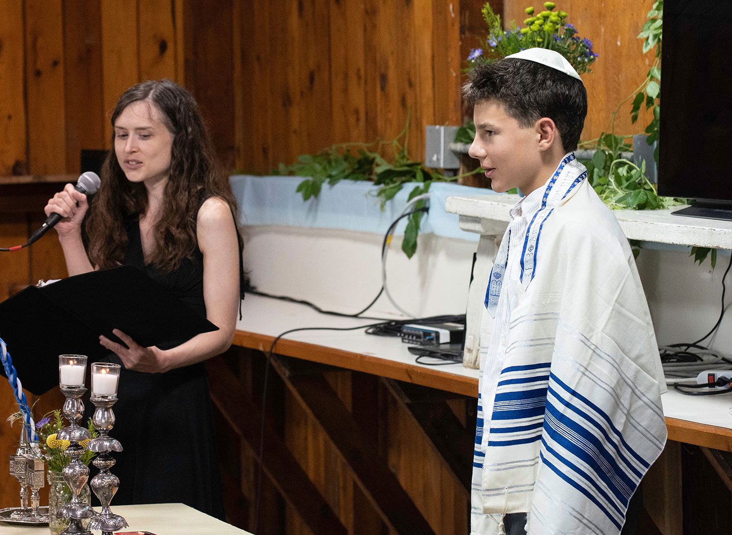 Rabbi Eva Cohen, wearing a black sleeveless dress, stands at a podium speaking into a microphone while looking toward Eli, a young B’Mitzvah celebrant. Eli stands to the right, dressed in a white button-down shirt and a blue and white striped tallit (prayer shawl). Two lit candles in silver holders are visible on the table in the foreground.