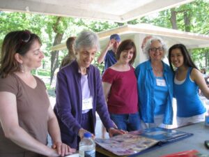 A group of women of various ages smiling and gathering around a table at an outdoor Or Emet event. They are looking at a large photo album together. The woman on the left is wearing a brown tank top, while others are in casual summer attire like red and blue shirts. Lush green trees and a white pavilion structure are visible in the background.