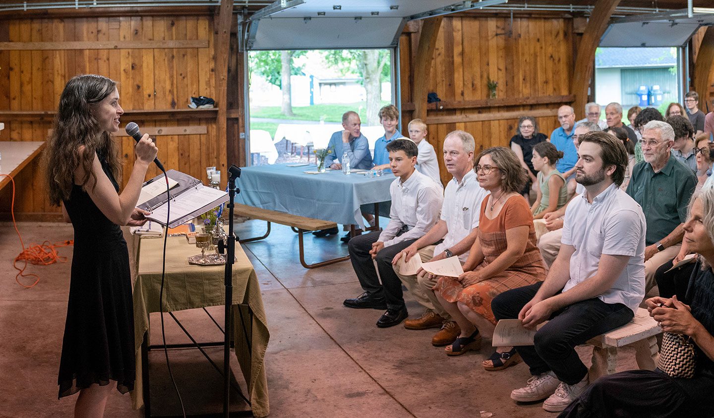 Rabbi Eva Cohen speaking into a microphone at a wooden podium during a B’Mitzvah ceremony. She is standing on the left in a black dress, looking toward a seated audience. The audience, including several adults and a young man in a white shirt, sits on wooden benches in a rustic, barn-like setting with large open doors revealing a green outdoor space.