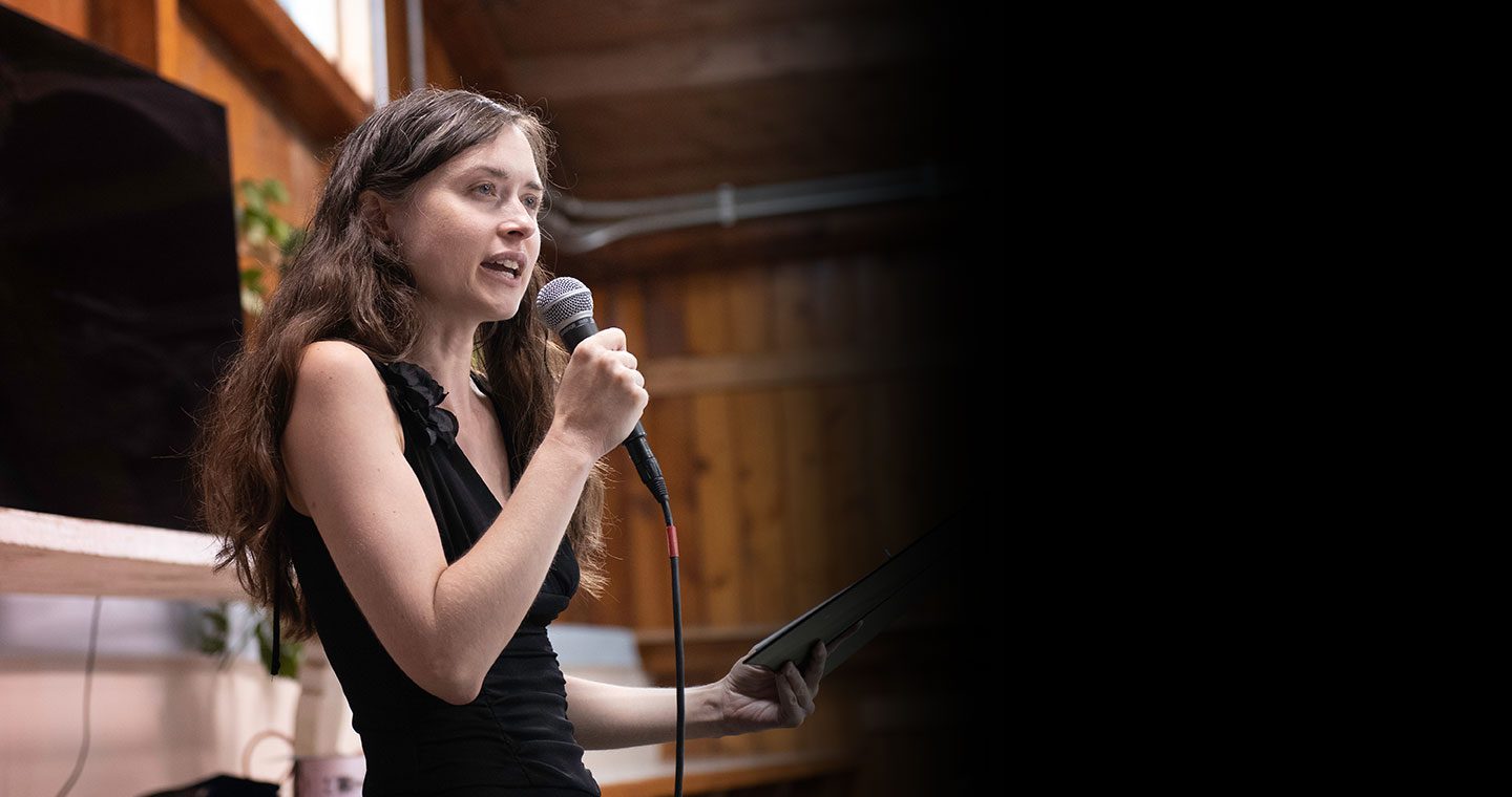 Rabbi Eva Cohen, with long dark hair and wearing a black sleeveless dress, speaking into a microphone during a B’Mitzvah ceremony. She is holding a tablet and looking toward the side with an engaged expression. The background features warm wood-paneled walls and a blurred indoor setting.