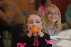 A young girl at an Or Emet event smiling while holding an orange slice in her mouth like a grin. Another girl and several adults are blurred in the background, seated at tables with plates of fruit during a Tu BiShvat seder celebration.