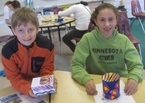 Two young students, a boy and a girl, smiling while sitting at a table in a classroom. They are proudly holding handmade, brightly decorated Tzedakah boxes. The boy on the left holds a rectangular box with colorful geometric patterns, and the girl on the right holds a cylindrical container painted with vibrant blue, yellow, and red floral designs.