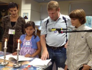 A man and two children standing at a table during a Shabbat service at Or Emet. The man is adjusting a microphone while a young girl in a blue cardigan looks down at a prayer book. Two lit white candles in silver holders are on the table in the foreground, and another woman stands behind them observing the ceremony.