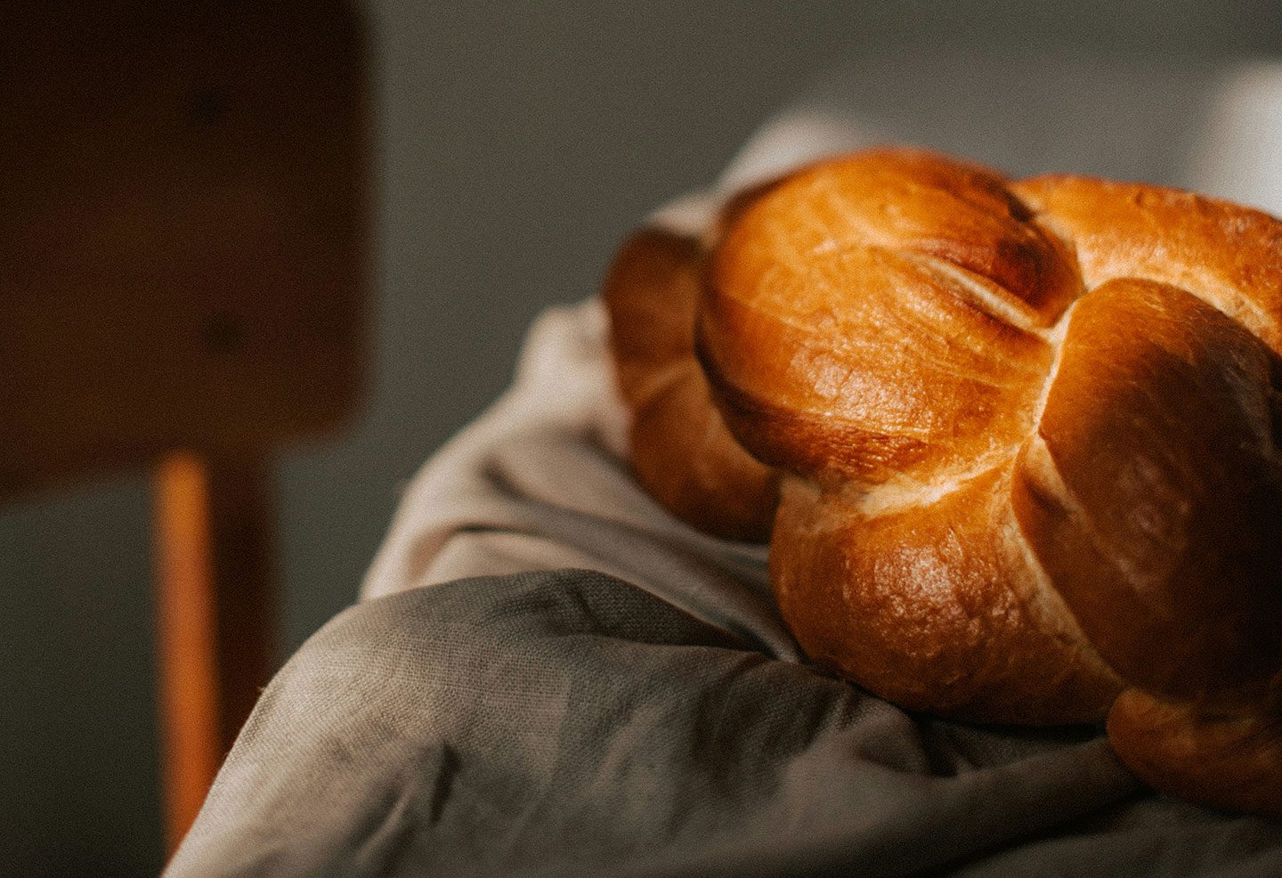 A close-up of a golden-brown, braided challah loaf resting on a soft, grey textured cloth. The bread has a glossy crust, highlighting the intricate braid, and is set against a warm, softly blurred indoor background.