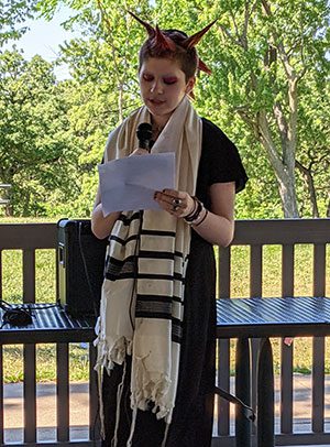 A young B’Mitzvah celebrant standing on a wooden deck outdoors, wearing a black outfit and a traditional tallit (prayer shawl). They are holding a piece of paper and speaking into a microphone during a ceremony, with lush green trees in the background.