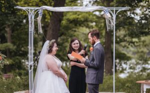 A bride and groom standing under a white metal chuppah during an outdoor wedding ceremony. The bride is wearing a white lace gown and a flower crown, while the groom is in a grey suit. An officiant stands between them holding a microphone and a small orange card. The background features lush green trees.