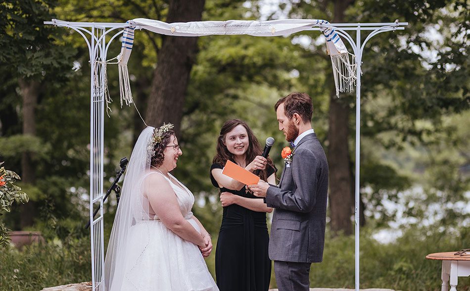 A bride and groom standing under a white metal chuppah during an outdoor wedding ceremony. The bride is wearing a white lace gown and a flower crown, while the groom is in a grey suit. An officiant stands between them holding a microphone and a small orange card. The background features lush green trees.