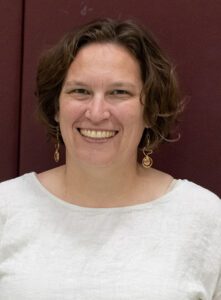 A portrait of Molly, a member of the Or Emet leadership team. She is a woman with shoulder-length wavy brown hair and glasses, wearing a white top and decorative gold earrings, smiling warmly against a dark red background.
