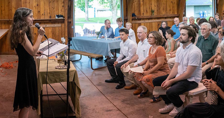 Rabbi Eva Cohen speaking into a microphone at a podium during an indoor Or Emet gathering. She is wearing a black sleeveless dress and looking toward a seated audience of diverse community members. The setting is a rustic room with wooden walls and large open doors leading to a green outdoor space.