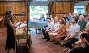 Rabbi Eva Cohen speaking into a microphone at a podium during an indoor Or Emet gathering. She is wearing a black sleeveless dress and looking toward a seated audience of diverse community members. The setting is a rustic room with wooden walls and large open doors leading to a green outdoor space.