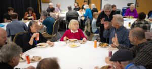 A group of Or Emet members gathered around a large white circular table for a Yom Kippur break-the-fast meal. People of various ages are eating, talking, and gesturing while seated in a social hall. Plates of food and cans of soda are on the table, and other occupied tables are visible in the background.