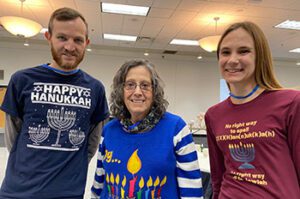 Three Or Emet members smiling together at a Hanukkah celebration. A man on the left wears a blue "Happy Hanukkah" t-shirt with a menorah, a woman in the center wears a blue and white striped sweater with a Hanukkah pin, and a young woman on the right wears a maroon shirt with a "Humanorah" logo and the words "No right way to be Jewish."