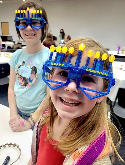 Two young girls smiling and wearing festive blue "Happy Chanukah" novelty glasses with yellow LED candle flames on top. The girl in the foreground is wearing a red shirt and a purple knit vest, while the girl behind her is in a light blue t-shirt.