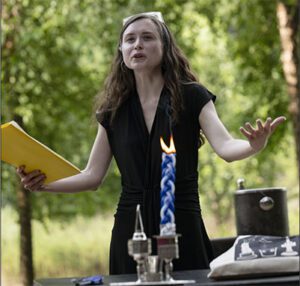 Rabbi Eva Cohen standing outdoors behind a table, gesturing with her hands while leading a ceremony. In front of her is a tall, lit blue-and-white braided Havdalah candle, a silver kiddush cup, and other ceremonial items. She is wearing a black dress and holding a yellow folder.