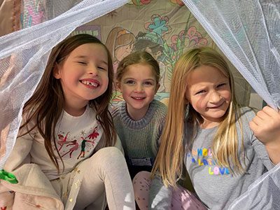 Three young girls smiling together under a mesh canopy during a Sukkot celebration. The girl on the left has long brown hair and is laughing, while the two girls next to her smile warmly for the camera. A colorful mural is visible in the background through the mesh.