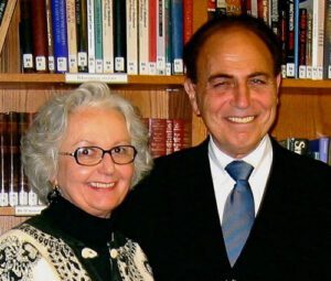 Rabbi Sherwin Wine, the founder of Humanistic Judaism, stands smiling in front of a bookshelf filled with books. He is wearing a dark suit with a light blue tie and is pictured alongside a woman with grey hair and glasses who is wearing a black and white patterned jacket.