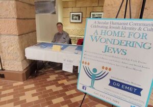 A close-up of an Or Emet promotional sign on an easel inside a building. The sign features a blue "Humanorah" logo and the text "A Secular Humanist Community Celebrating Jewish Identity & Culture: A HOME FOR WONDERING JEWS." In the background, a man sits at a table with a blue tablecloth and community materials.