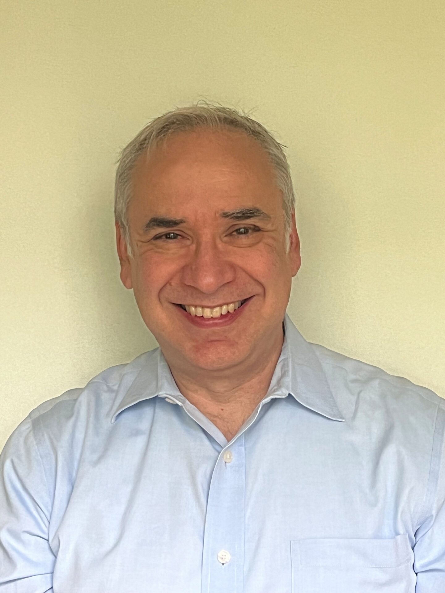 A professional headshot of Sergei, a middle-aged man with short grey hair and a warm smile. He is wearing a light blue button-down shirt and is positioned against a neutral, light-colored background.