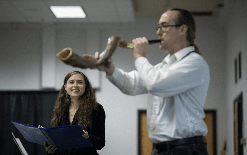 Rabbinic candidate Eva Cohen stands on the left holding an open book, smiling as she watches Dan Gladen blow a large shofar. They are indoors at the Minnesota JCC Sabes Center during a Yom Kippur service.