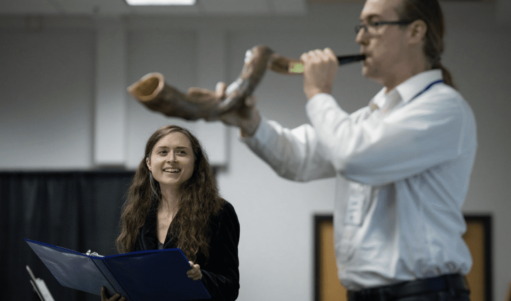 Rabbinic candidate Eva Cohen stands on the left holding an open book, smiling as she watches Dan Gladen blow a large shofar. They are indoors at the Minnesota JCC Sabes Center during a Yom Kippur service.