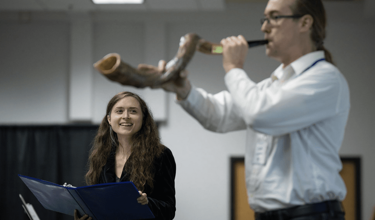 Rabbinic candidate Eva Cohen stands on the left holding an open book, smiling as she watches Dan Gladen blow a large shofar. They are indoors at the Minnesota JCC Sabes Center during a Yom Kippur service.