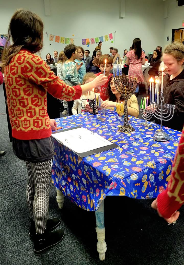 A group of children and adults gathered around a table with a blue Hanukkah-themed tablecloth, lighting several silver menorahs. A young girl in a red patterned sweater and checkered leggings stands in the foreground, watching the candles being lit in a brightly lit community room.